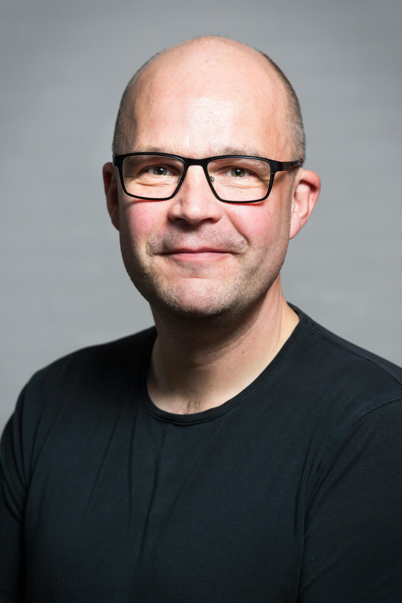 Portrait of a smiling bald man wearing glasses and a black t-shirt against a neutral background.