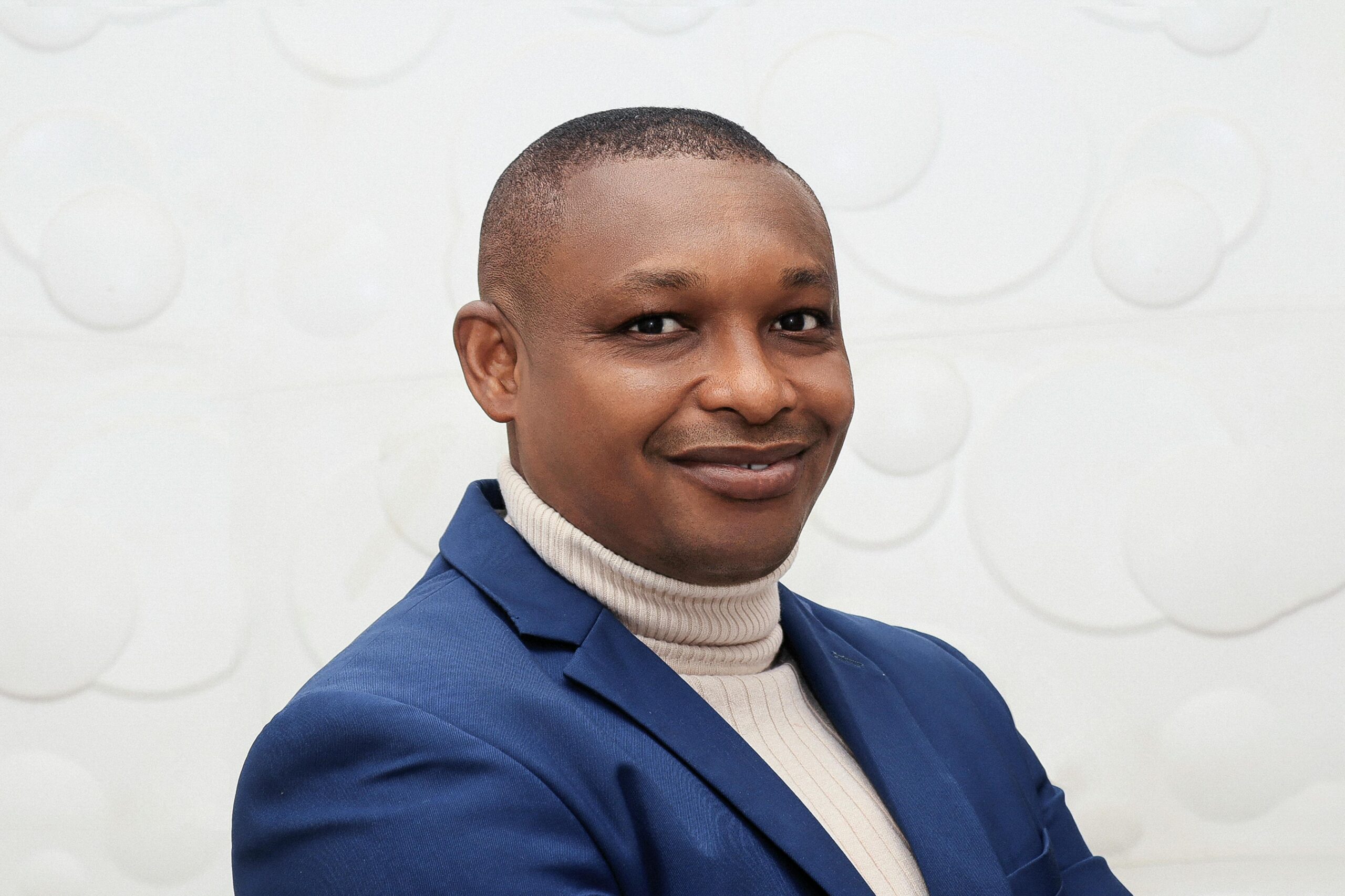Portrait of a smiling man in a blue suit and turtleneck against a white background.