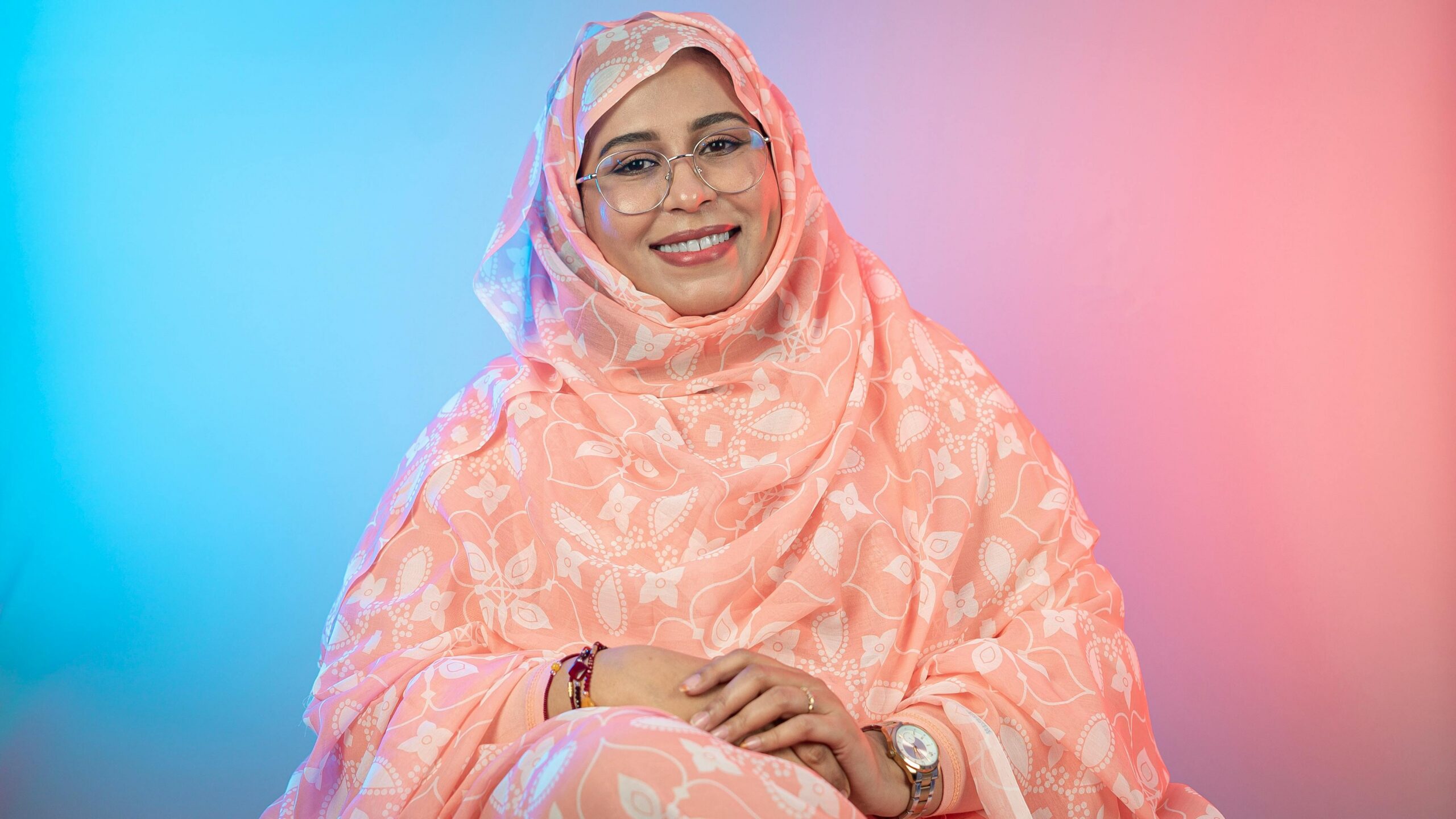 Smiling woman in traditional peach floral hijab with pastel background, studio shot.