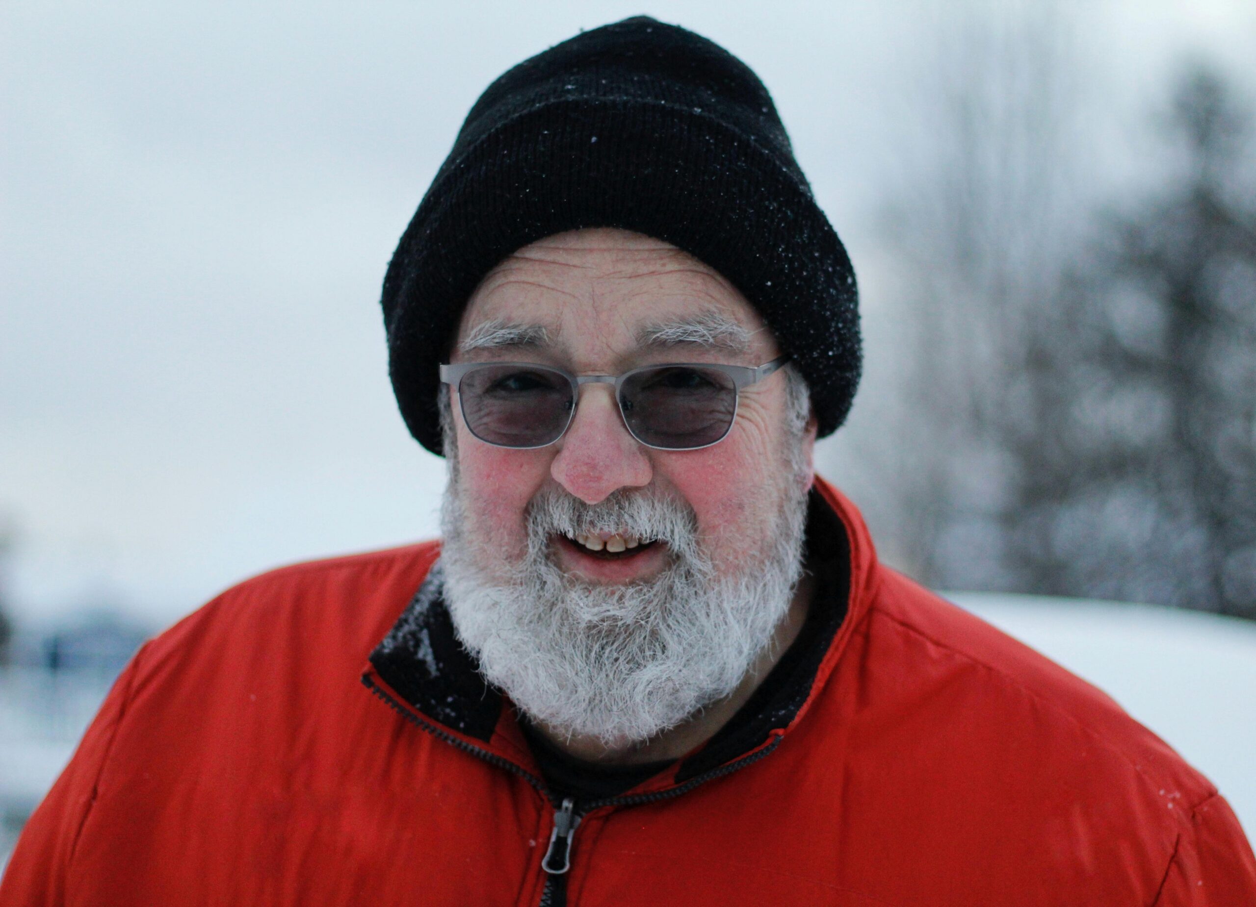 An elderly man with a white beard smiles warmly while wearing a red coat and black beanie in a snowy outdoor setting.