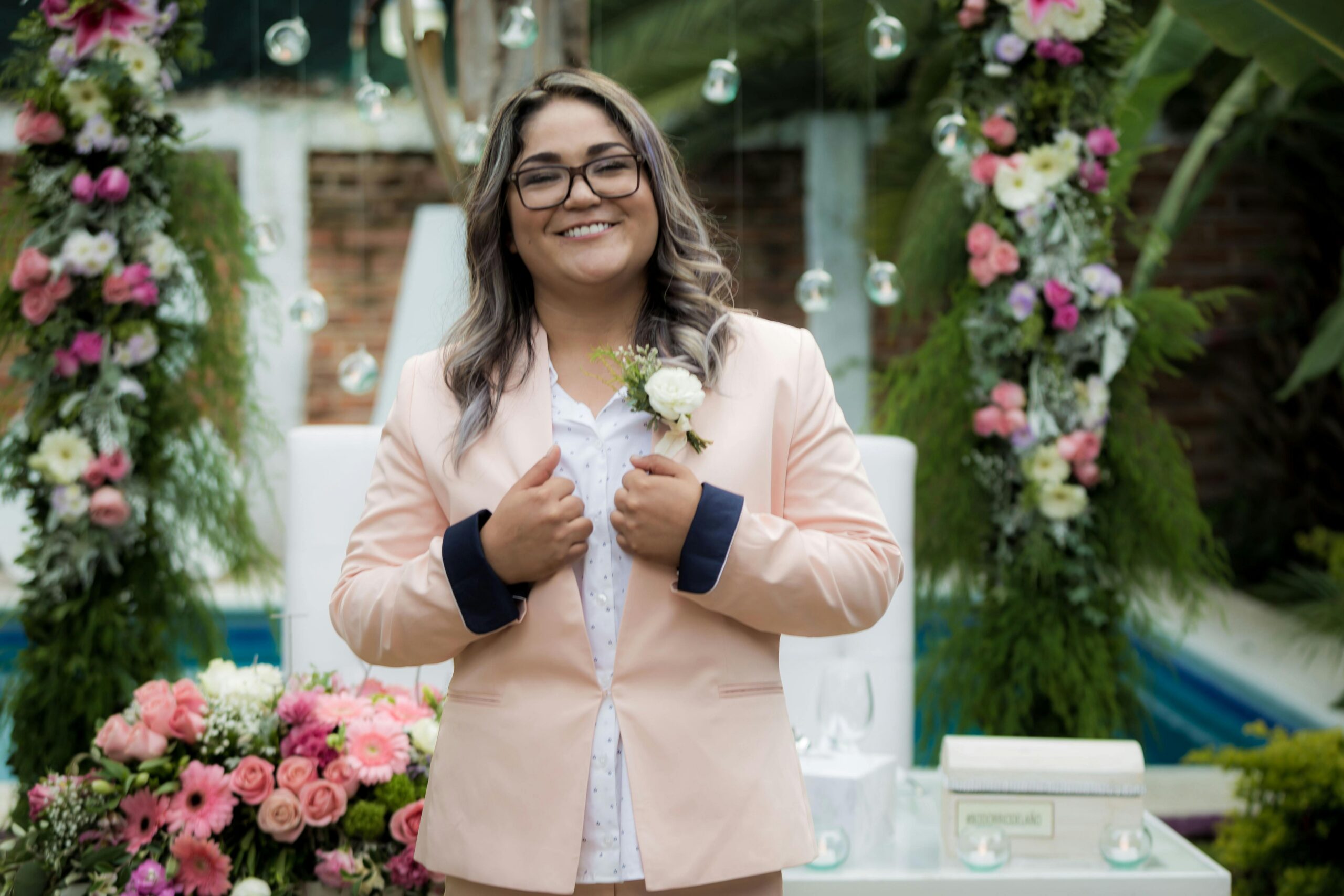 Smiling woman in peach blazer posing confidently at outdoor wedding surrounded by flowers.