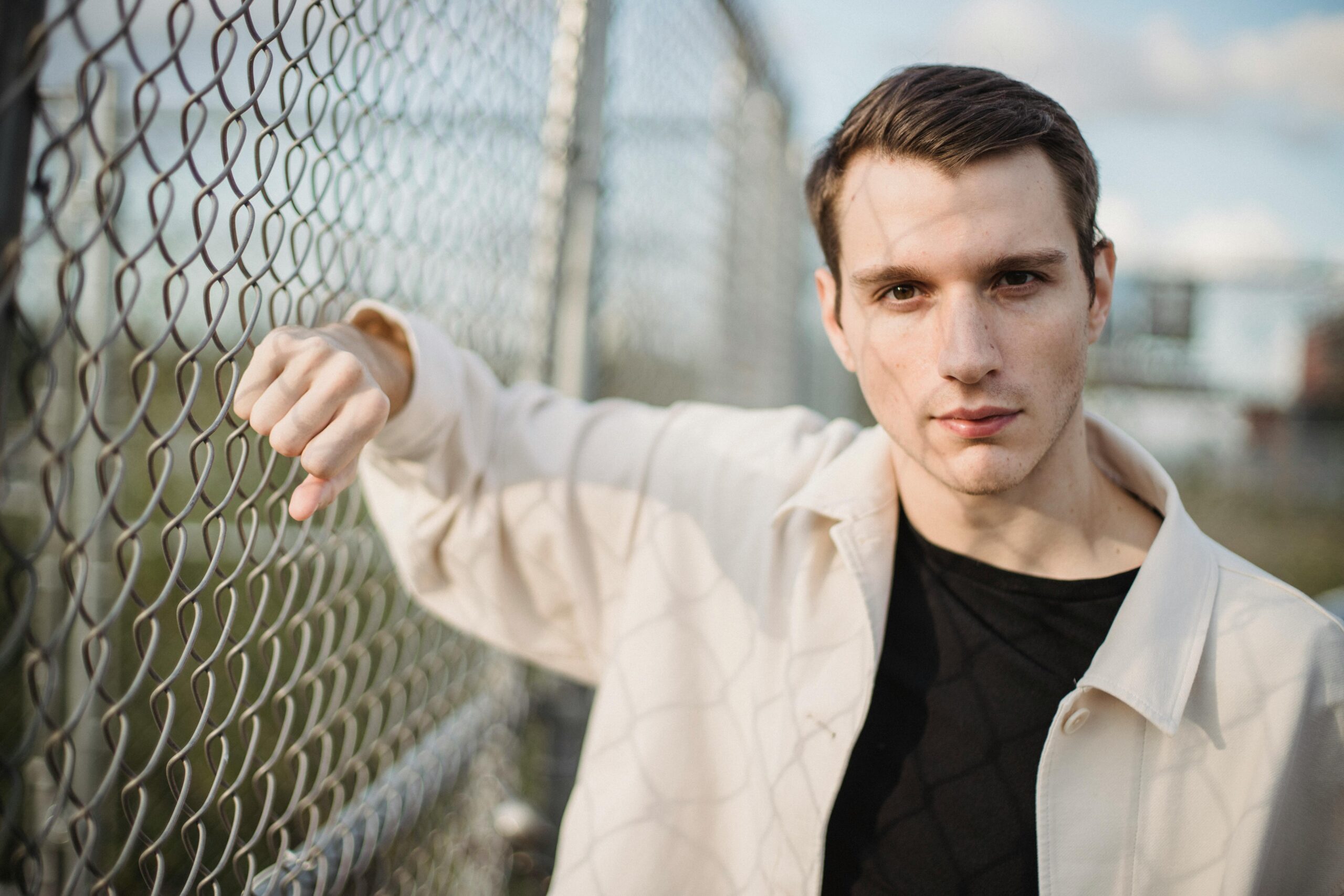 Young serious male in shadow wearing casual outfit leaning on metal net and looking at camera