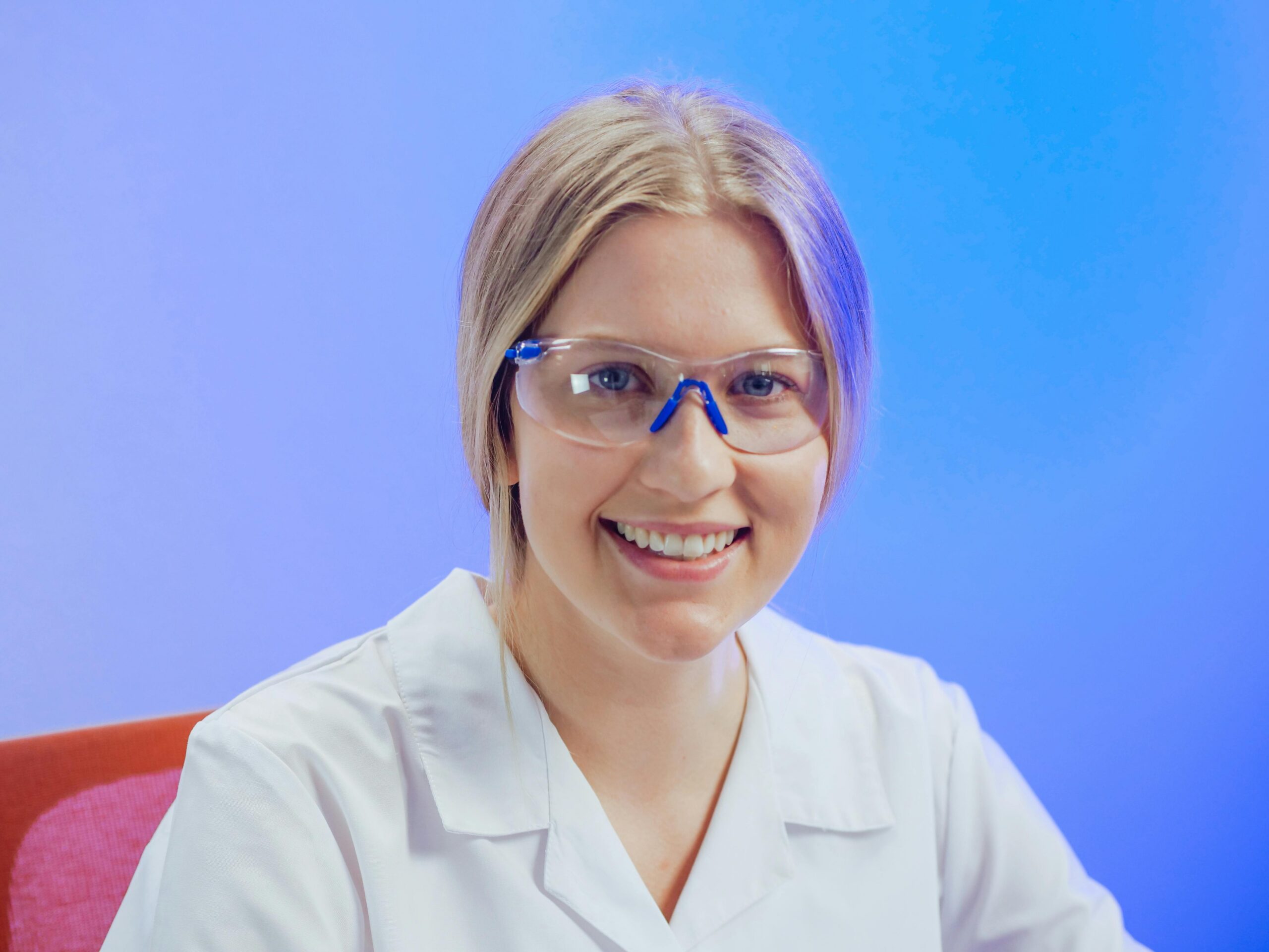 A cheerful female scientist wearing safety goggles and a lab coat smiling in a lab setting.